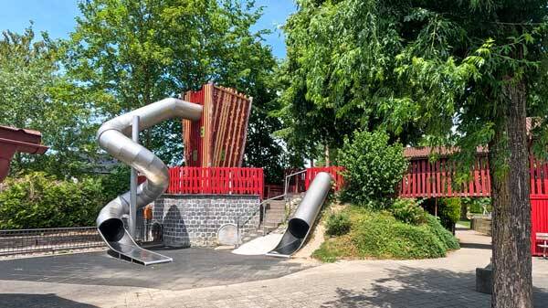 Moderner Kinderspielplatz mit einer grossen silbernen Tunnelrutsche, einer Hangrutsche und hölzernen Klettertürmen.