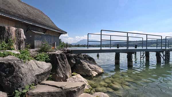 Ein moderner Steg mit Metallgeländer ragt neben einem alten Holzhaus in das klare Wasser des Sempachersees, umgeben von felsigem Ufer.