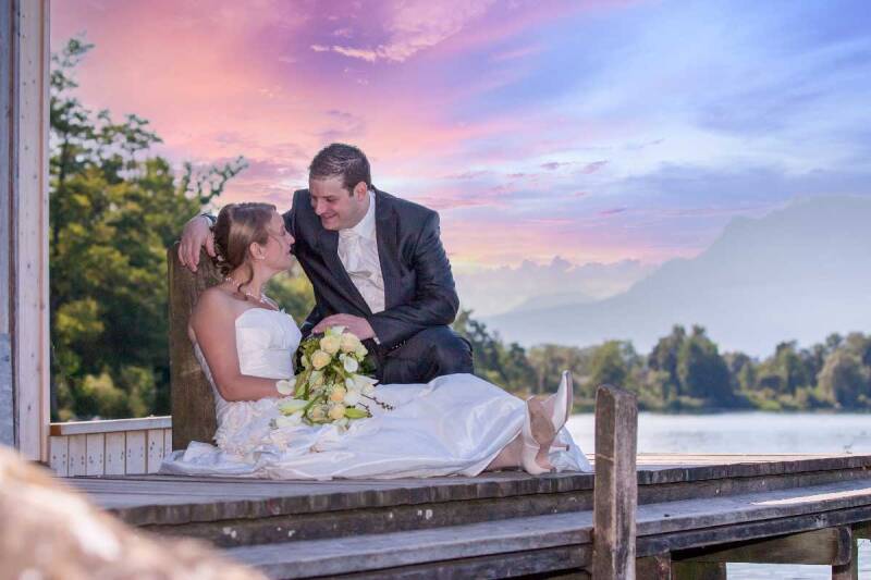 Brautpaar sitzt verliebt auf einem Holzsteg am See mit Bergen im Hintergrund bei Sonnenuntergang