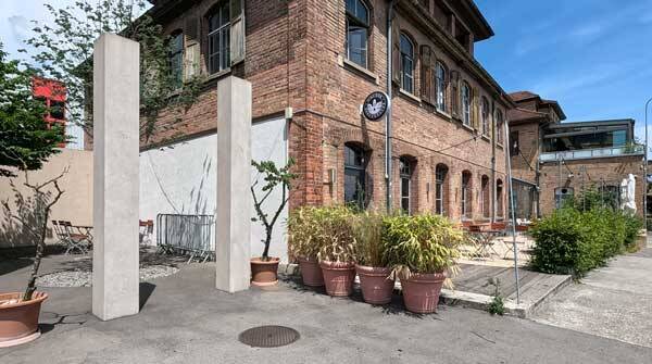 Historisches Industriegebäude aus Backstein mit grossen Fenstern, davor Pflanzkübel und ein Aussenbereich mit Tischen.