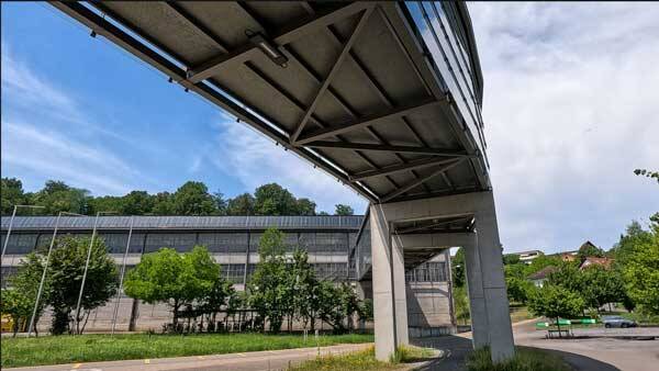 Blick von unten auf eine moderne, überdachte Fussgängerbrücke aus Beton und Metall vor einem grossen Industriegebäude.