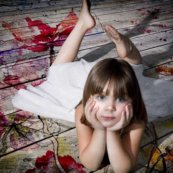 Ein kleines Mädchen liegt auf dem Boden und schaut mit abgestütztem Kinn direkt und neugierig in die Kamera.