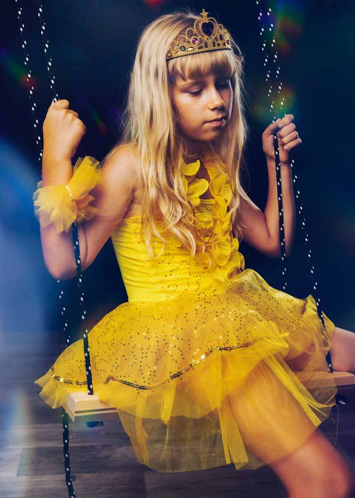 Blondes Mädchen mit goldener Krone und gelbem glitzernden Prinzessinnenkleid sitzt träumend auf einer Schaukel im Studio