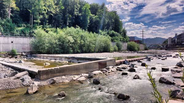 Detailaufnahme der Kleinen Emme mit Betonbauten zur Flussregulierung und grossen Steinen im strömenden Wasser, im Hintergrund ein bewaldeter Hügel.