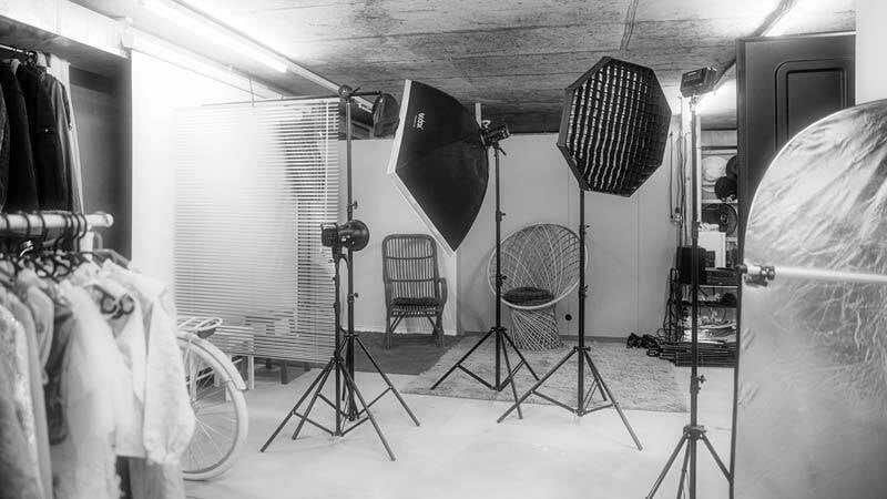 Portraitbereich im Fotostudio mit verschiedenen Studioblitzen, einem stilvollen Korbsessel und Kleiderstangen mit Outfits.