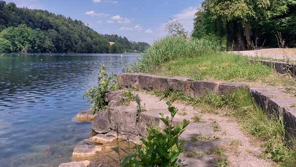 Nahaufnahme der terrassenförmigen Steinstufen am Ufer des Rotsees mit grünem Schilf und Blick auf das ruhige Wasser und die gegenüberliegenden bewaldeten Hänge.