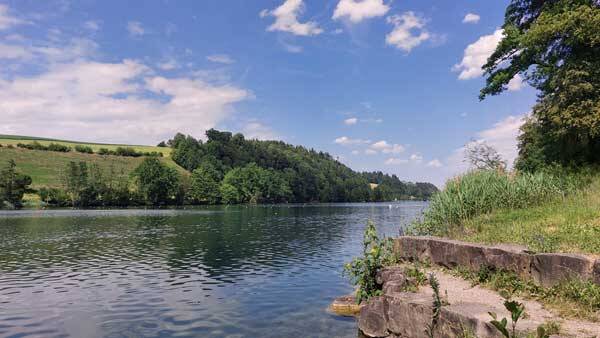 Weiter Blick über den ruhigen Rotsee bei Luzern mit bewaldeten Hügeln und grünen Feldern im Hintergrund sowie einer steinernen Uferböschung im Vordergrund.