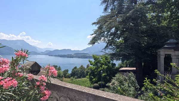 Blick vom Park über rosa Blüten auf den See und Berge.