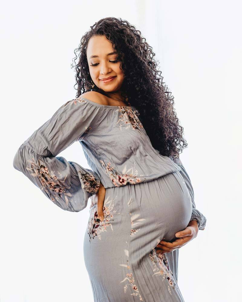 Eine hell belichtete Aufnahme einer lächelnden schwangeren Frau mit langen Locken. Sie trägt ein hellblaues Off-Shoulder-Kleid mit Blumenmuster und blickt sanft nach unten auf ihren Babybauch.