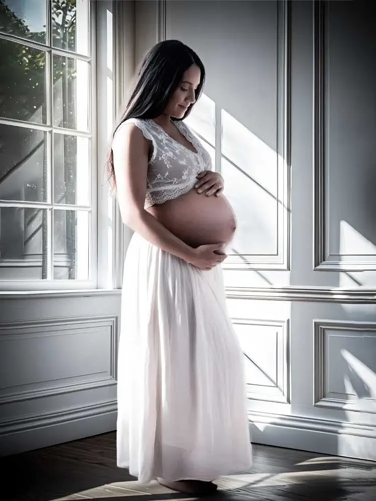 Helles Babybauch-Fotoshooting am Fenster: Schwangere Frau in weissem Spitzenkleid und fliessendem Rock.