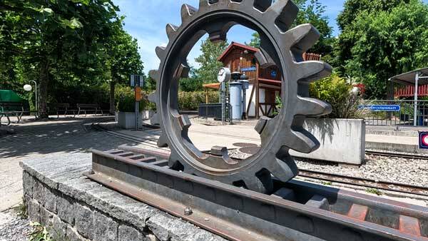 Ein grosses, eisernes Zahnrad als Skulptur auf Schienen, im Hintergrund eine kleine hölzerne Bahnstation unter blauem Himmel.