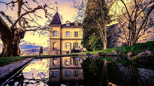 Eine historische Villa mit Turm unterhalb der Museggmauer in Luzern, deren Fassade sich perfekt in einem ruhigen Teich im Vordergrund spiegelt, umgeben von alten Bäumen vor einer abendlichen Bergkulisse.