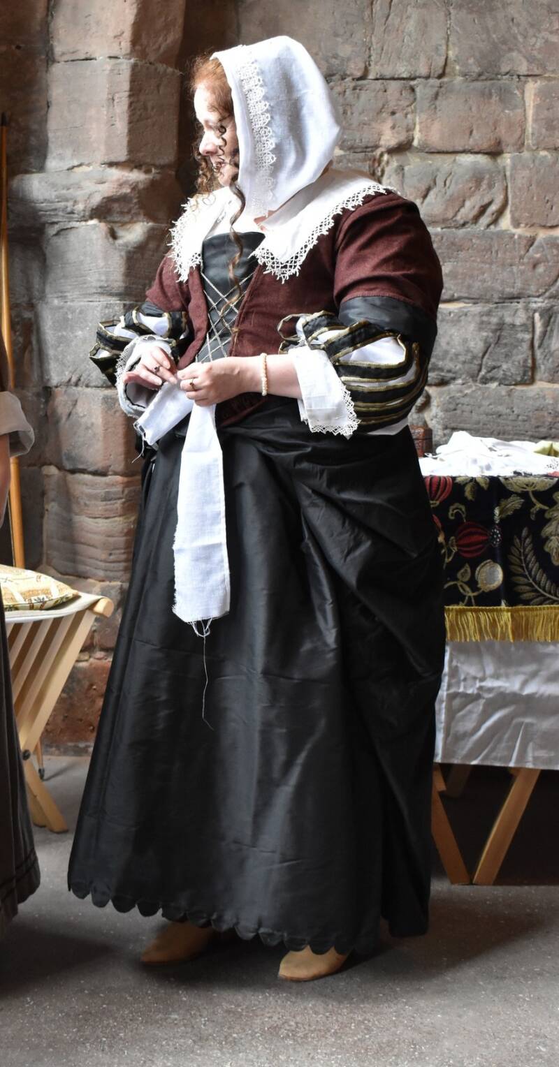 Photo of a woman dressed in high status clothing from the 1640s. She wears a wine coloured gown over a black silk petticoat. She has lace edged collar, cuffs and loose hood.  Rings adorn her fingers and pearl bracelets around her wrists