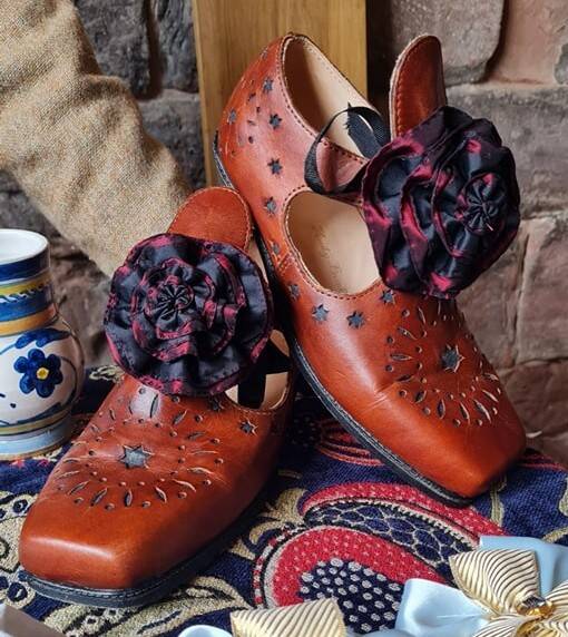 Image of a pair of replica mid 17th Century shoes. Made from reddish brown leather, with punched decoration in the form of stars, the shoes have square toes and are fastened with rosettes made from balck and dark red satin