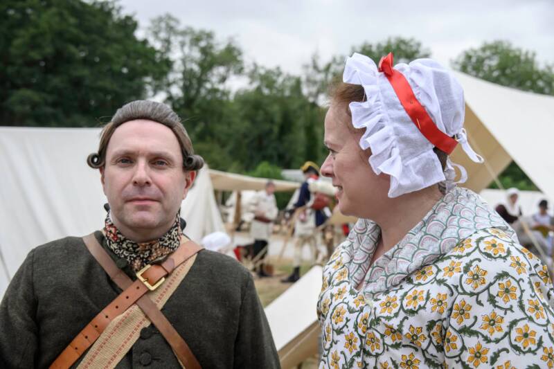 Photo of a man and woman (head and shoulders) dressed in late 18th Century fashions. The man wears a brown wig with rolls of hair above each ear, and a dark wool coat. The woman wears a white frillled cap and a white gown with yellow flowers