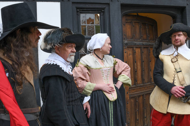Photo of thre men and a woman all dressed in clothing from the mid 17th Century. They stand outside a half timbered, black and white house. A wooden door with iron studs stands ajar behind them