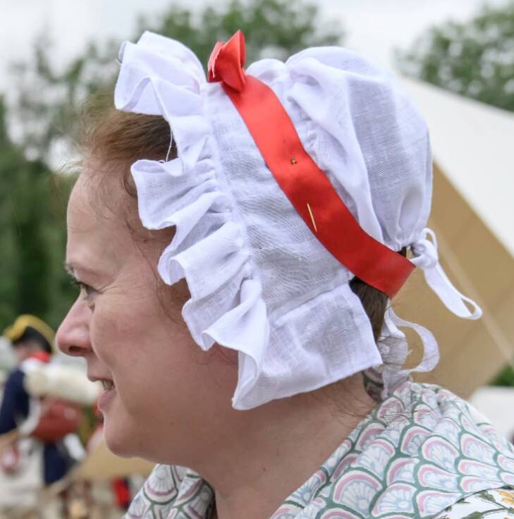 Photo of a woman's head in profile. She has brown hair brushed straight back from her forehead. The rest of her hair is covered with a white linen cap with a frill trimming its front edge. The cap is secured to the head with a red ribbon tied in a bow. 