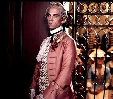 Photo of a man wearing a grey 18th Century wig. He wears a coat of pink satin with a pale coloured brocade waistcoat underneath, and a lace cravat