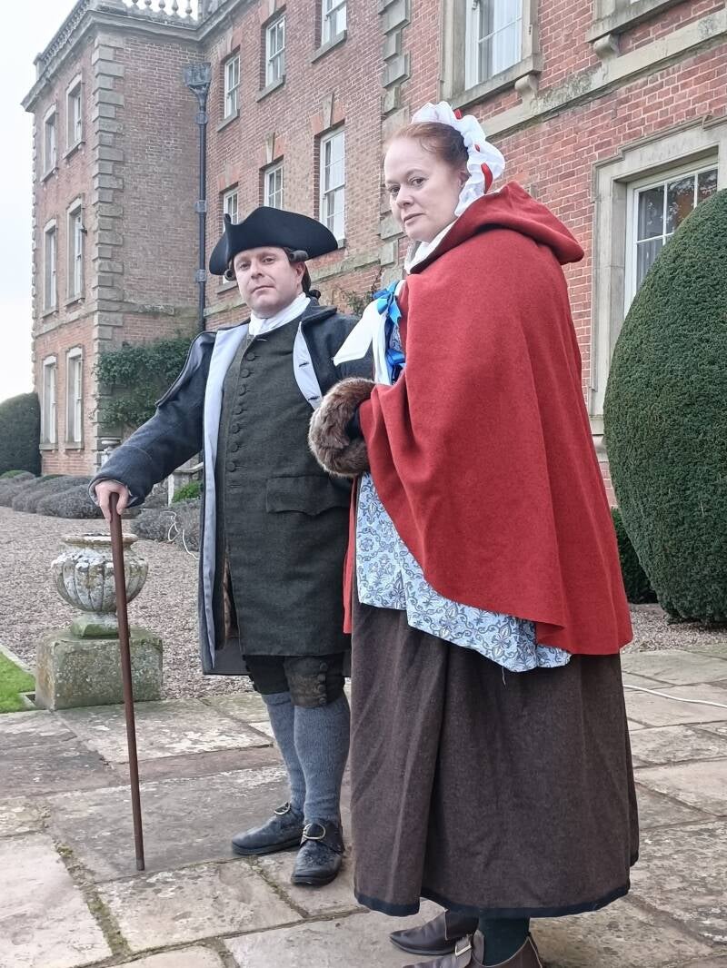 Photo of a man and woman dressed in 18th Century winter clothing, standing in front of a stately home. The man wears a grey great coat and black tricorn hat and the woman wears a red hooded cloak and carries a fur muff
