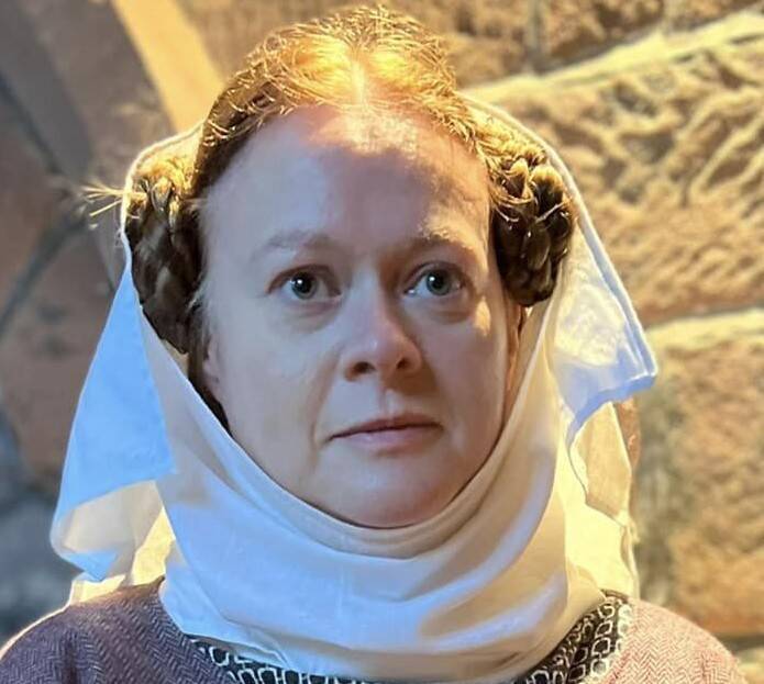 Photo of a woman's head. She has brown hair, middle parted and worn in two plaited buns on each side of her head. These are partially covered by a white linen wimple worn under the chin, and a matching short veil worn over the top of the head.