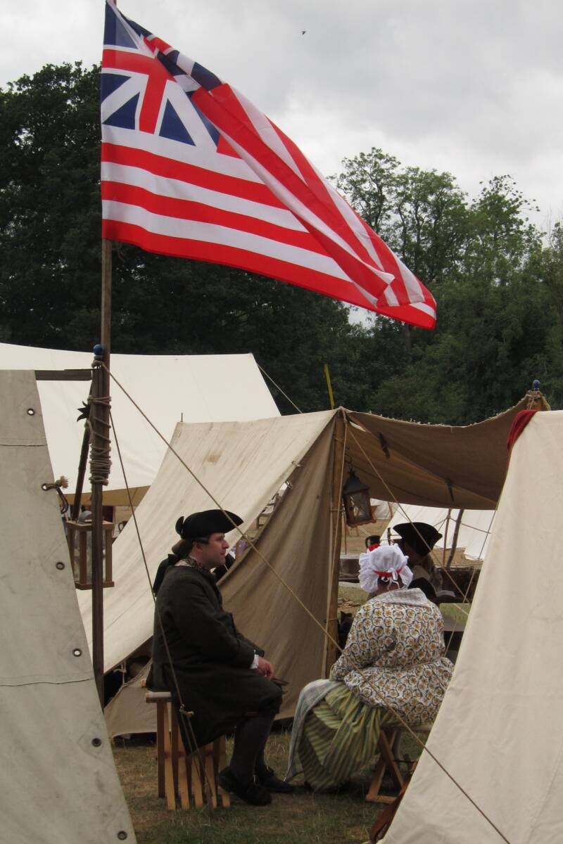 Photo of a man and woman wearing 18th Century clothing. They sit on folding stools amongst several canvas ridge tents. An American flag flutters above their heads