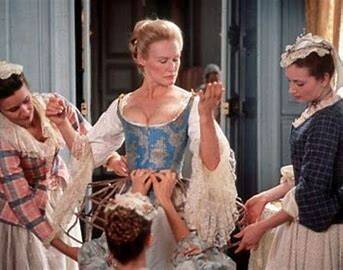 Photo of a woman being dressed in 18th Century clothing by three maids. The woman wears blue brocade corset, white undergarment with lace frills on the sleeves. She has fair hair worn pinned up.