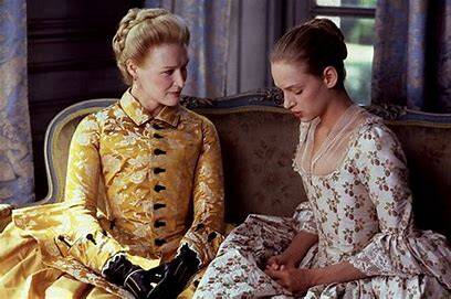 Photo of two women sitting on a settee wearing 18th Century clothing. Both women have fair hair pinned up. One woman wears yellow brocade, the other wears pale silk with small floral motifs