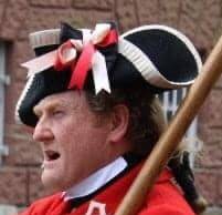 Photo of a man's head - clean shaven - with red hair tied back in a ponytail, wearing  a black cocked hat decorated with white braid and red and white ribbons.