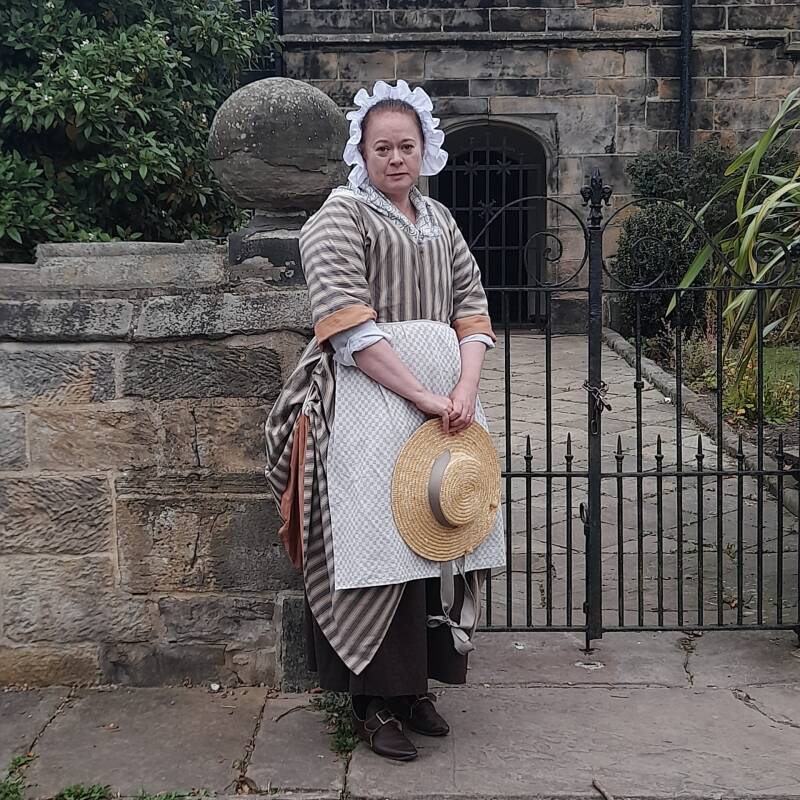 Photo of a woman standing infront of a gateway to a stone building behind. She is dressed in 18th Century fashions, including a white frilled cap, brown striped gown and holds a wide brimmed straw hat.