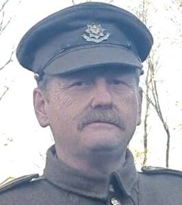 Photo of a man's head with a sandy red moustache. He wears a khaki cap with a peak and matching jacket