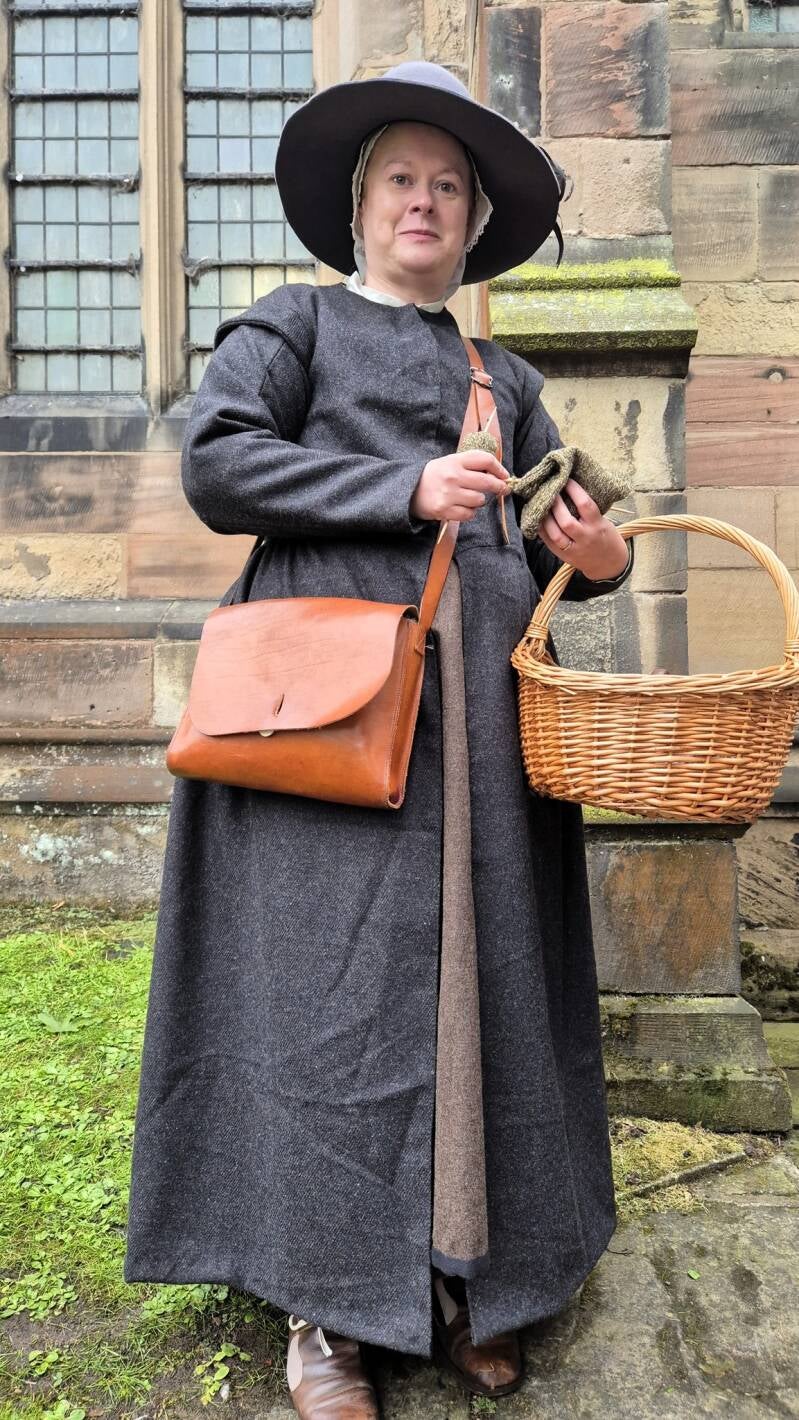 Photo of a woman wearing lower class clothing from the mid 17th Century. She wears a long dark grey gown over a brown petticoat, has a wide brimmed dark hat and carries a wicker basket and brown leather satchel type bag