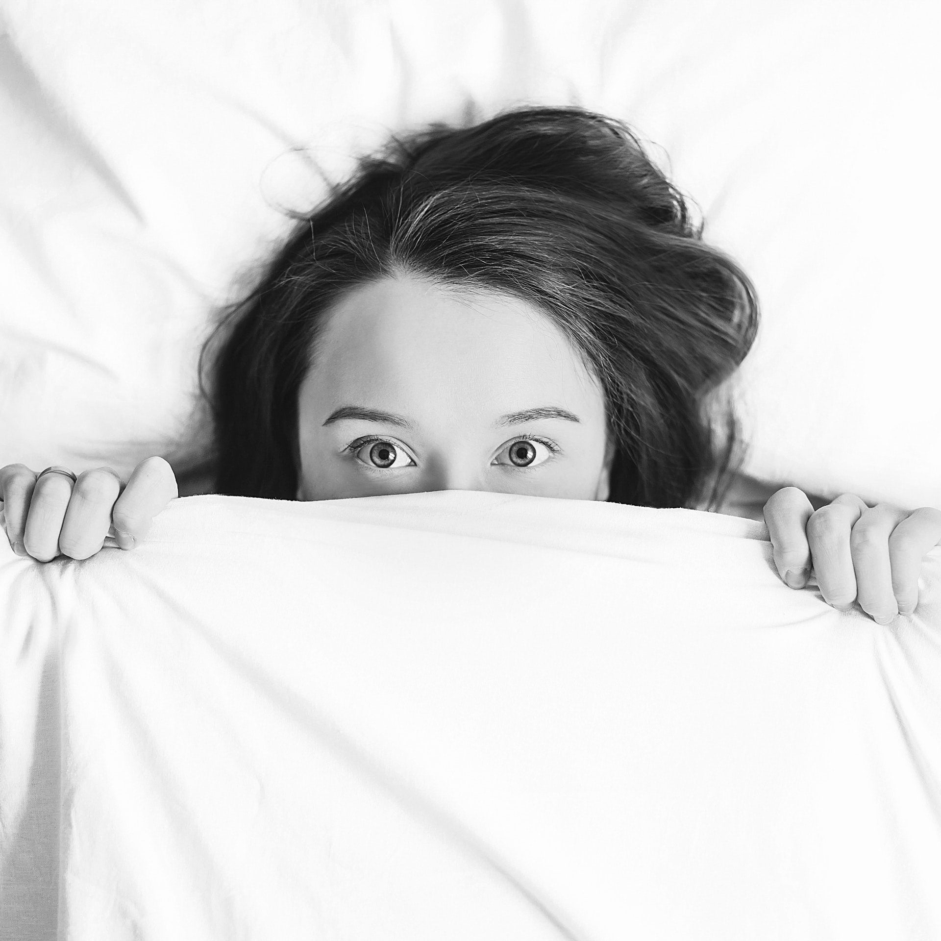 Femme allongée dans son lit, les yeux grands ouverts et le visage partiellement caché sous la couverture, illustrant les troubles du sommeil et l’insomnie apaisés par la méditation.
