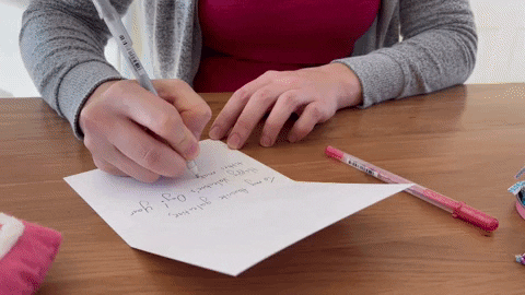  “A person writing a handwritten message inside a greeting card on a wooden desk with colourful chocolates nearby.”