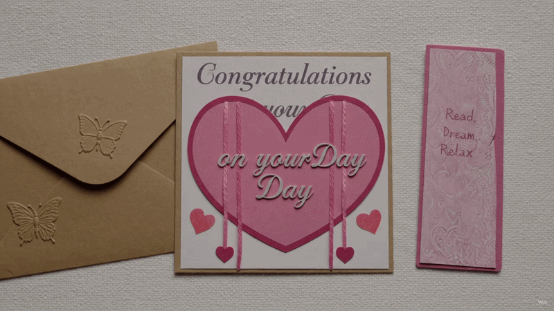 Handmade wedding card with pink heart design and matching bookmark, displayed with a brown envelope on a soft textured background.