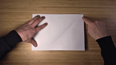 Hands folding a sheet of white paper on a wooden desk, showing the start of a handmade craft project