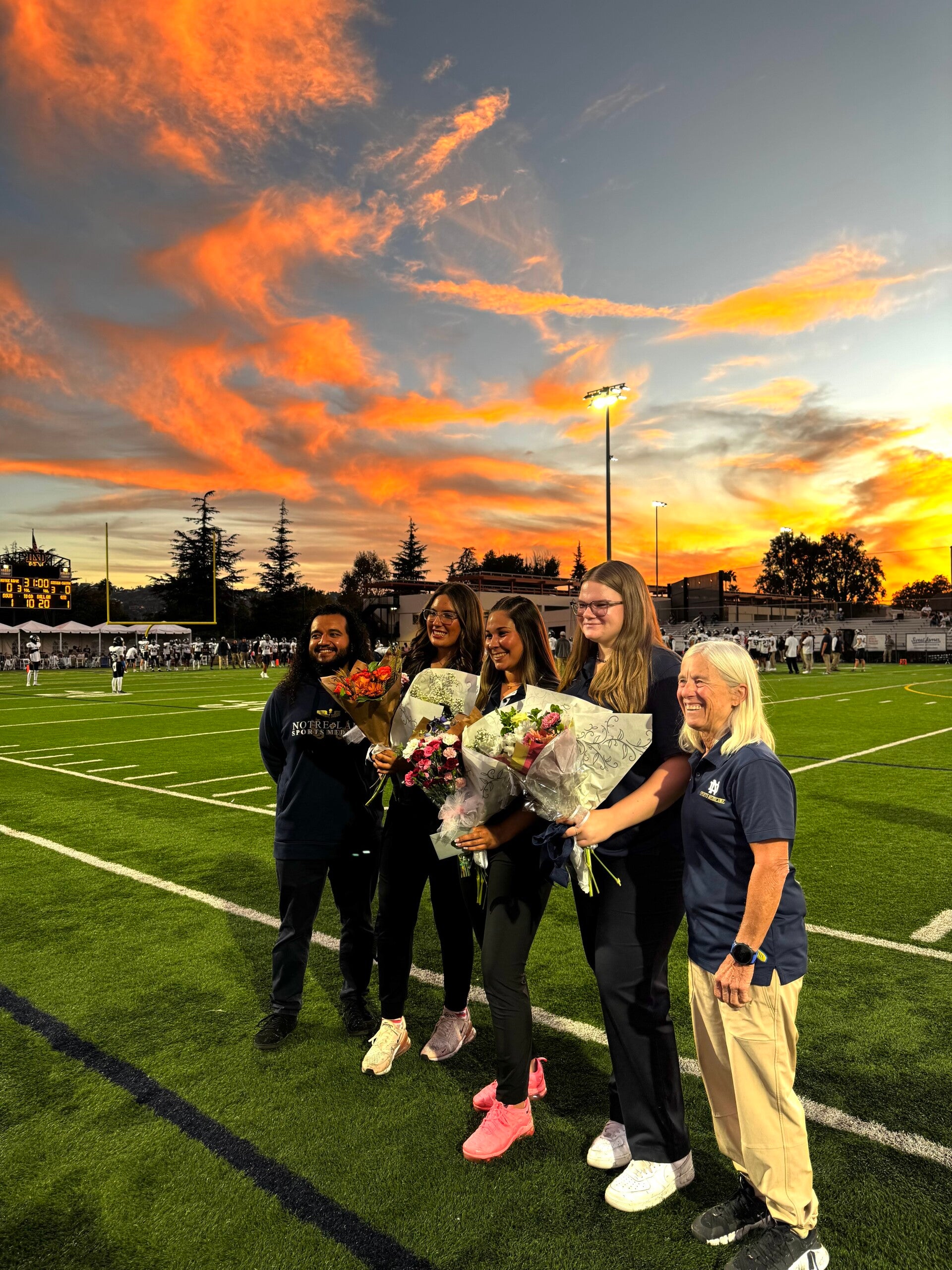 Holding flowers with 2 other friends and the head athletic trainers smiling on the football field posing for a picture on senior night. 