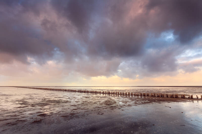 Wijds wadlandschap met een houten palenrij en weerspiegelingen onder een kleurrijke wolkenlucht.