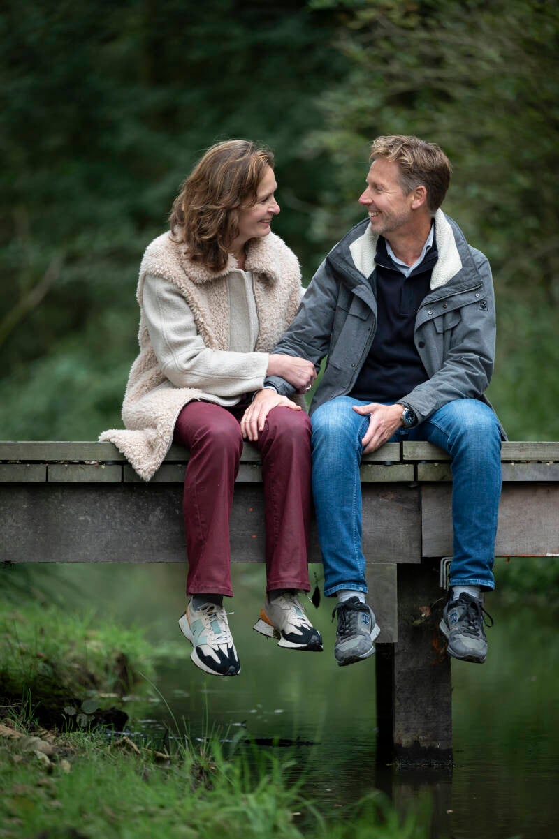 Twee mensen zitten naast elkaar op een houten bruggetje in het groen en kijken glimlachend naar elkaar.