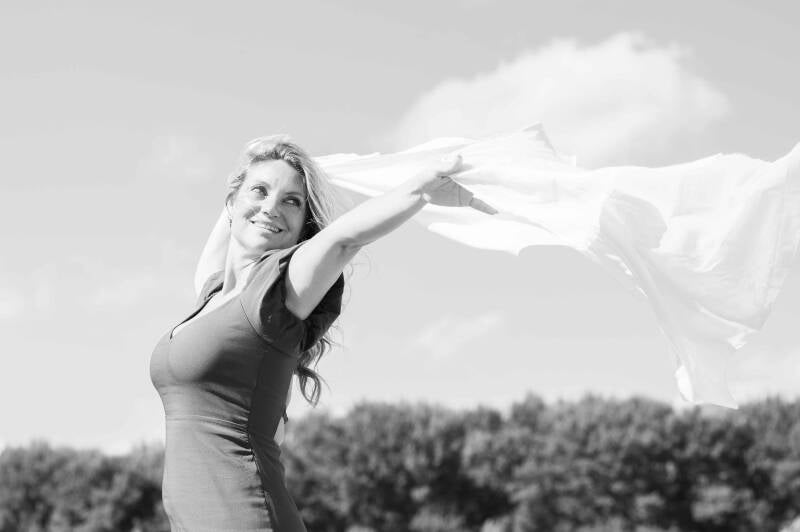 Zwart-witfoto van een vrouw die buiten glimlacht met een wapperende sjaal in haar uitgestrekte arm, met lucht en bomen op de achtergrond.