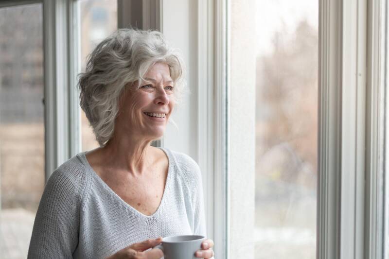 Femme mature aux cheveux gris souriante près d'une fenêtre, une tasse à la main.