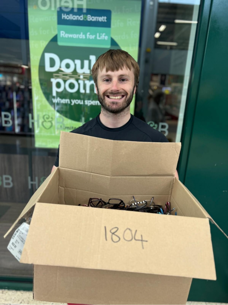 Lion Cobi stands outside an opticians shop with a lkarge box of spectacles.