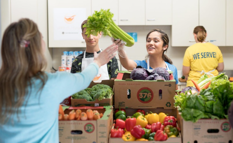 Photographic image of a mixture of male and femal lions Club members working in a community kitchen or food bank, wearing Lions clothing, and one with "we serve" written on the back.