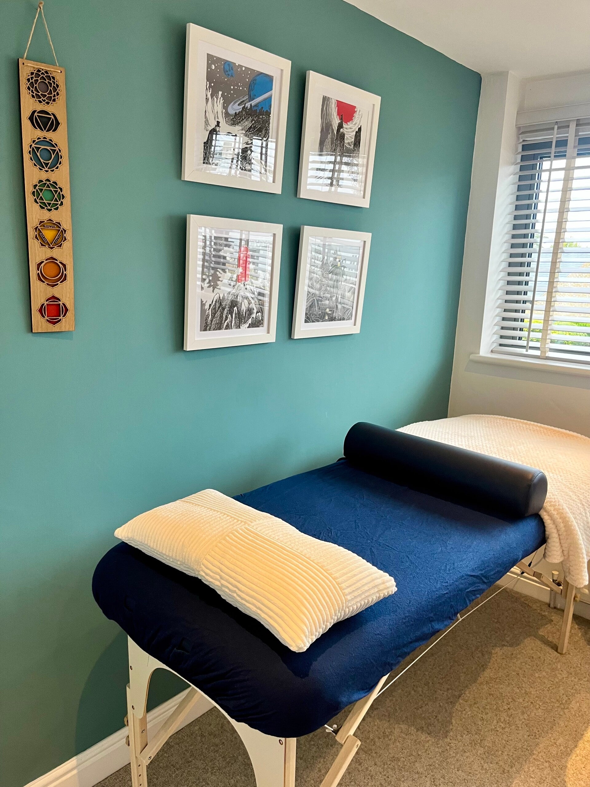 A treatment table with a soft pillow and blanket in a St Neots healing space, with framed prints and a chakra hanging on a teal wall