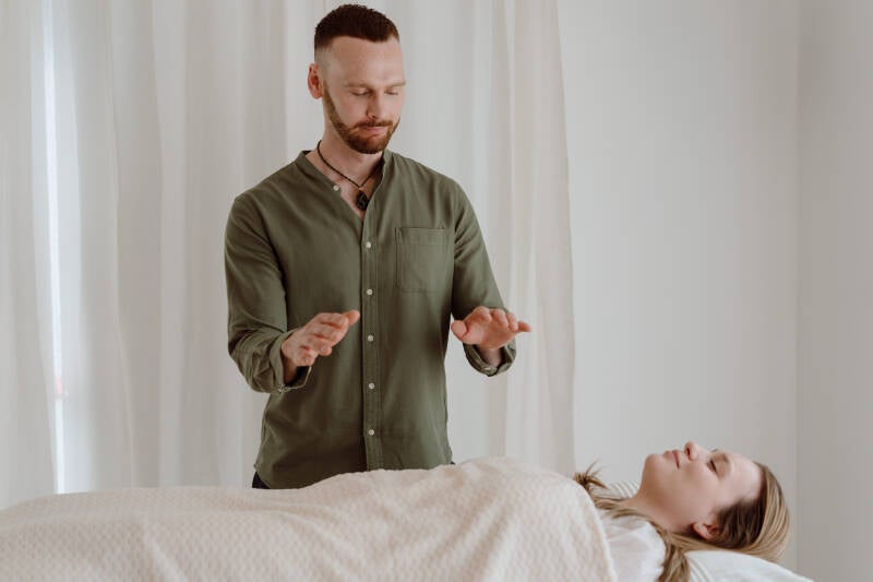 A Reiki practitioner standing beside a treatment table with his hands held above a client resting under a blanket