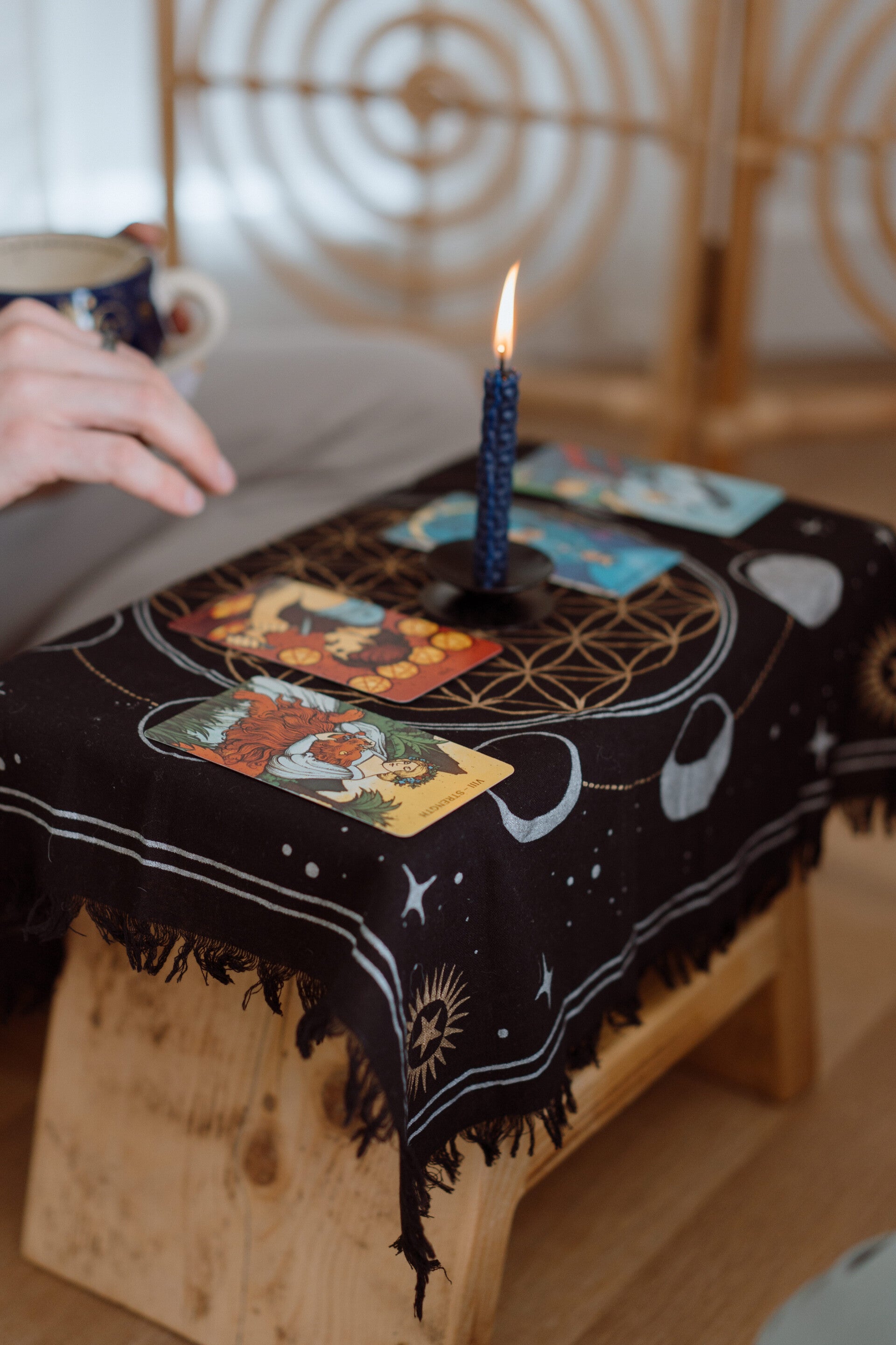 Tarot cards laid out on a black star-patterned cloth beside a lit blue candle, with a hand resting nearby holding a mug