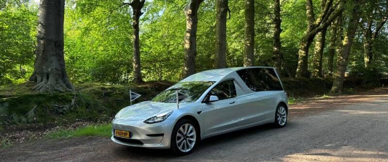 Een zilveren, elektrische rouwauto (Tesla) met rouwvlaggetjes, geparkeerd op een zandpad in een groene, bosrijke omgeving.