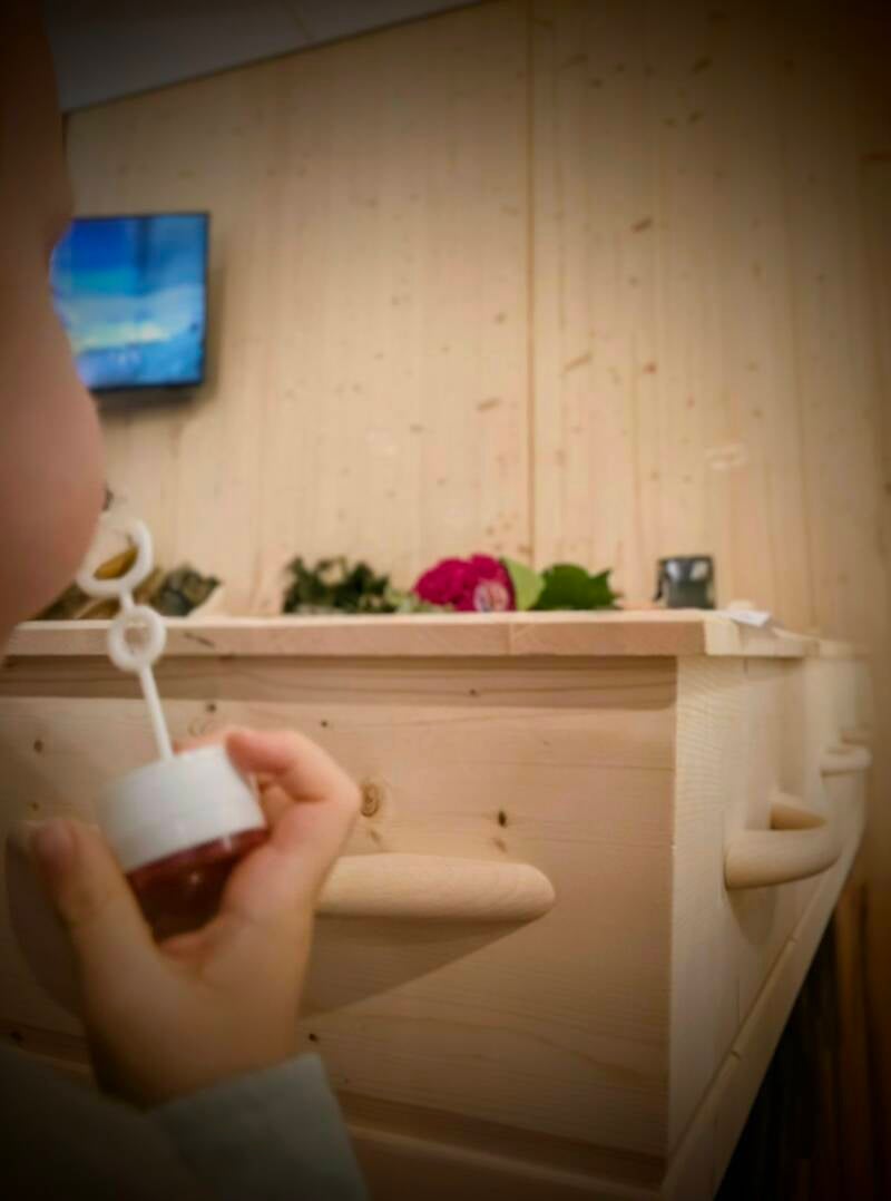 Een kind blaast bellenblaas naast een lichte houten grafkist waar bloemen op liggen.