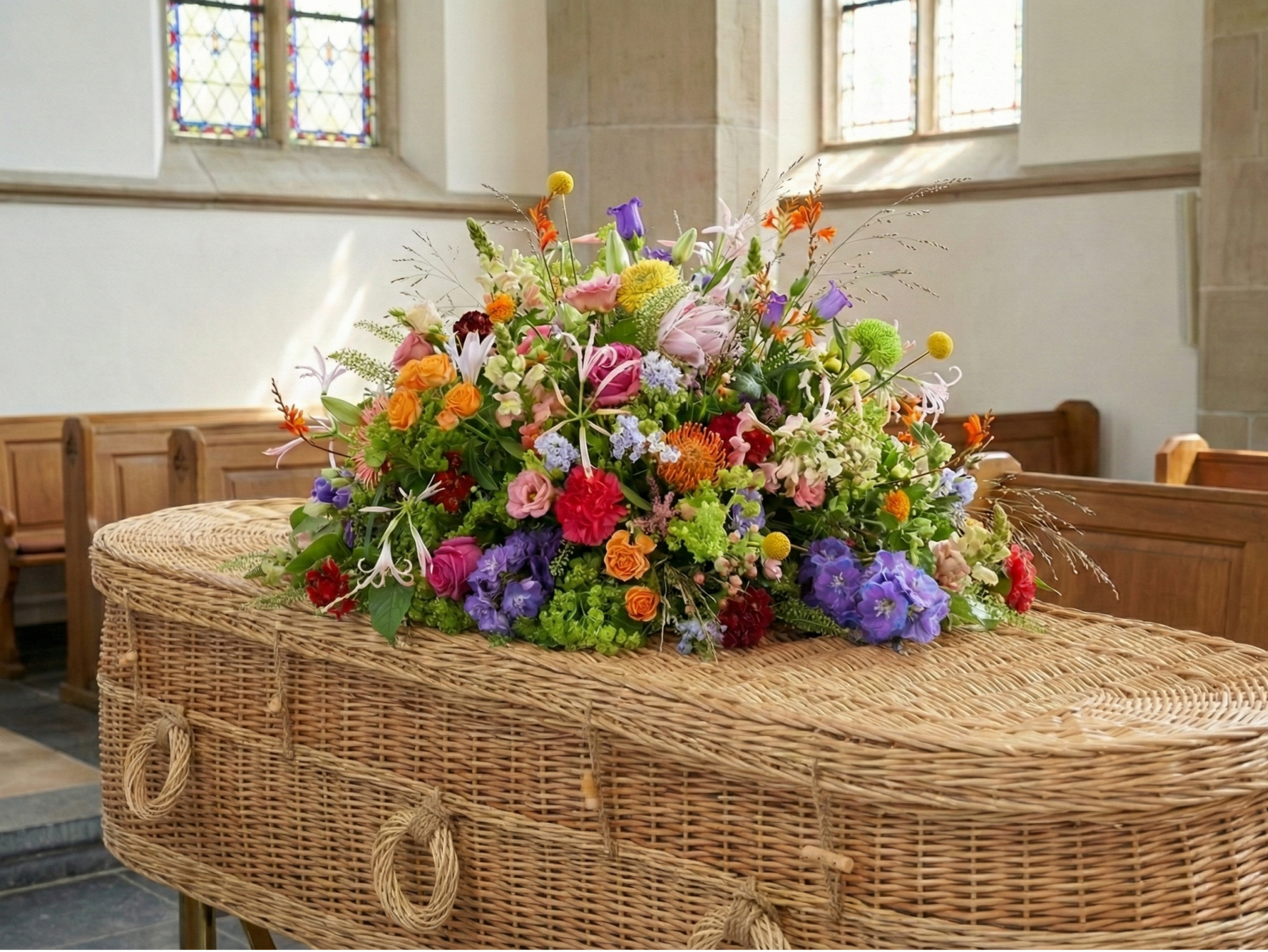 Een rieten uitvaartkist in een kerk of kapel met houten banken op de achtergrond. Op de kist ligt een groot, kleurrijk rouwboeket met diverse bloemen.