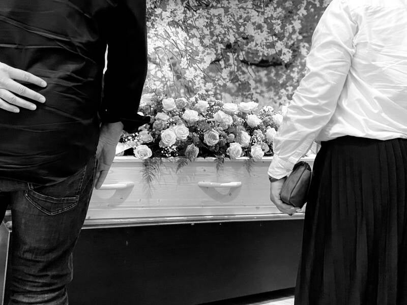 Een zwart-witfoto van nabestaanden die naast een uitvaartkist staan. De kist is versierd met een groot bloemstuk van rozen.