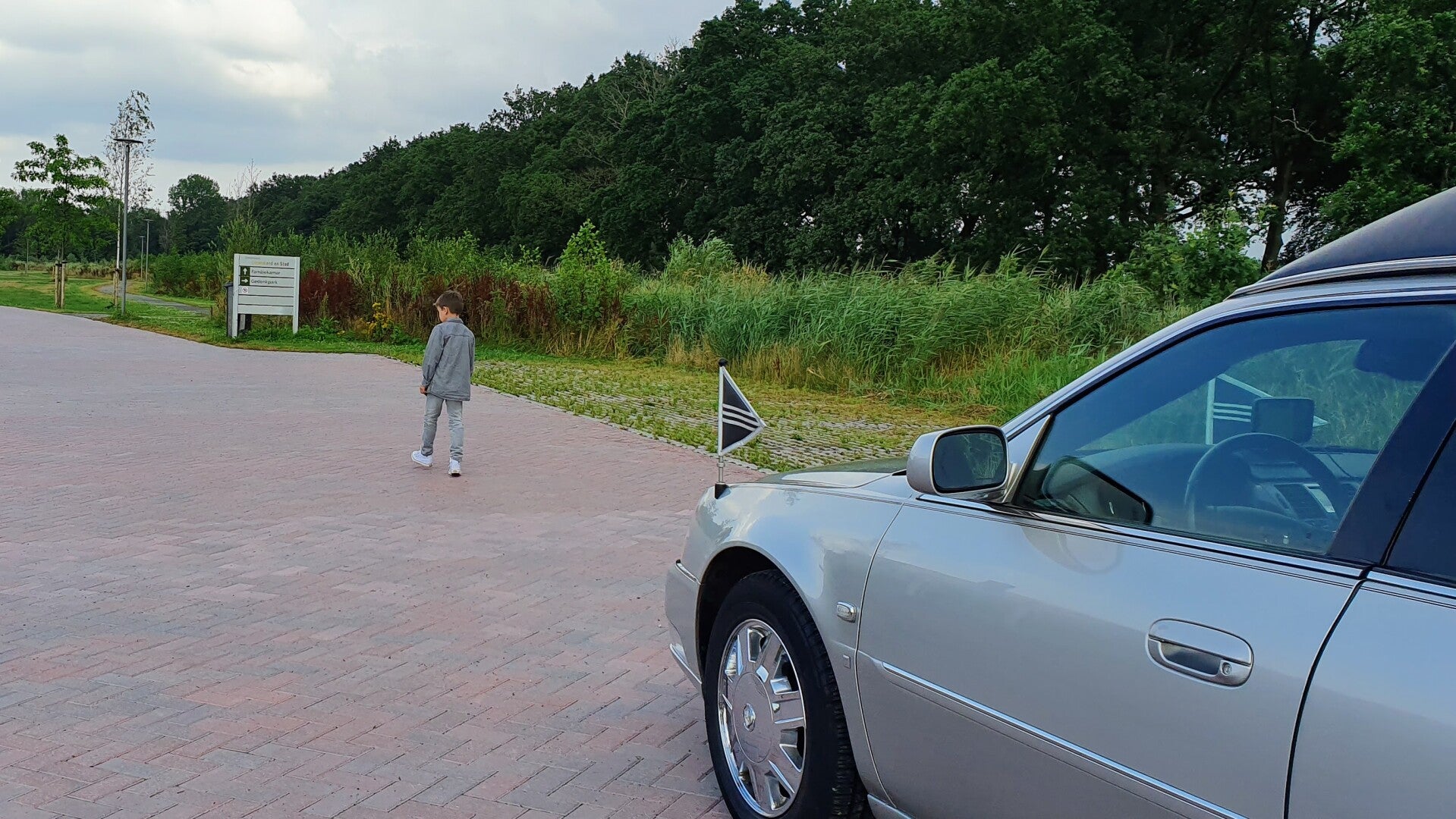 Een jongetje loopt op een pad in een groene omgeving nabij een geparkeerde zilveren rouwauto. Advies over kinderen betrekken bij een uitvaart door Jouw Afscheid.